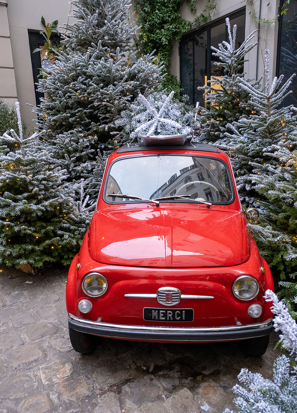 Small red car with 'Merci' license plate parked among snow-covered trees.