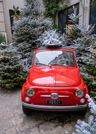 Small red car with 'Merci' license plate parked among snow-covered trees.